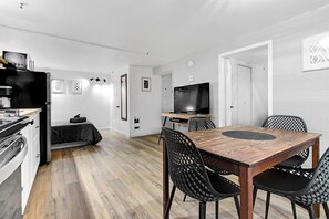 Eine moderne Küche mit integriertem Backofen, eine Essecke mit Holztisch und schwarzen Stühlen sowie ein Fernseher im Hintergrund.