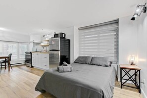 Ein modernes Schlafzimmer mit einem großen Bett, einem kleinen Schreibtisch, einer Küchenzeile und einem Fenster mit Jalousien.