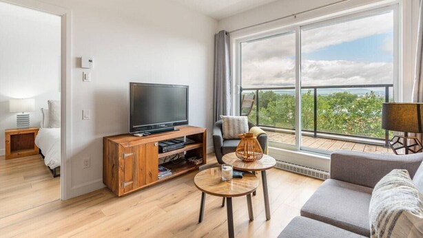 A modern living room with a flat-screen TV on a wooden stand, a sofa, a small round table, and a balcony with a view. - Cozy & Co | The GH View | Centre-ville + Parking (Québec City)