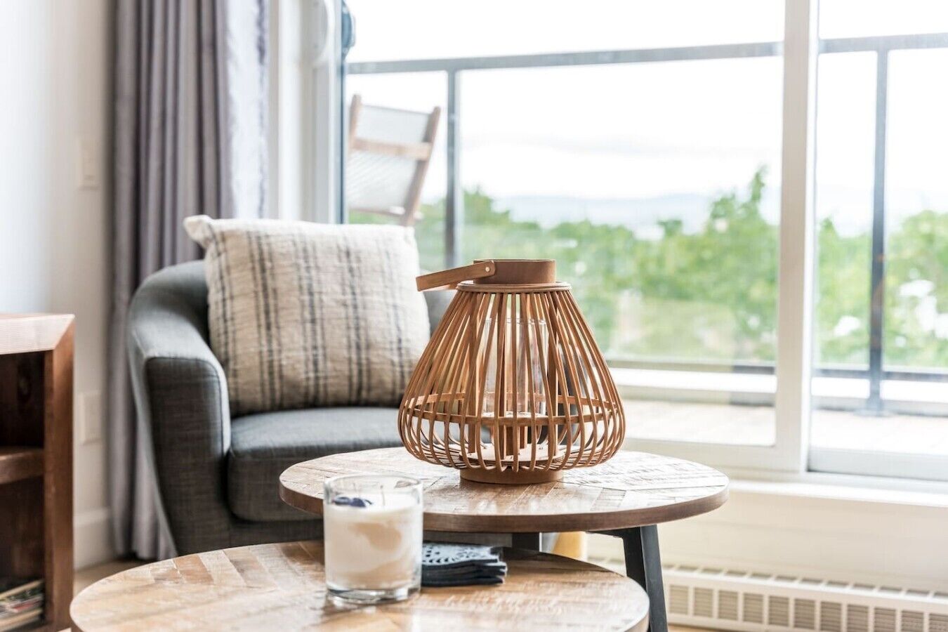 Une pièce avec une table ronde en bois, un chandelier, un verre de lait, un canapé gris avec un coussin et une vue sur la verdure par une fenêtre.