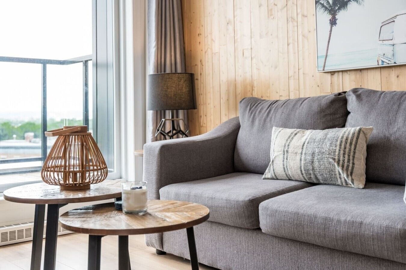 A modern living room with a grey sofa, a round wooden table, a lamp, and a window with a view.