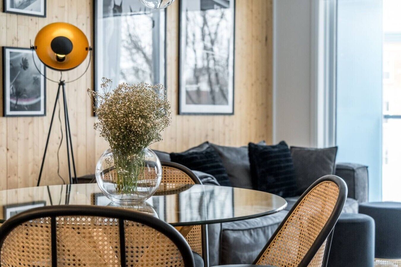 Un intérieur moderne avec une table ronde en verre, des chaises en osier, un vase de fleurs, une lampe sur pied et un canapé avec des coussins.