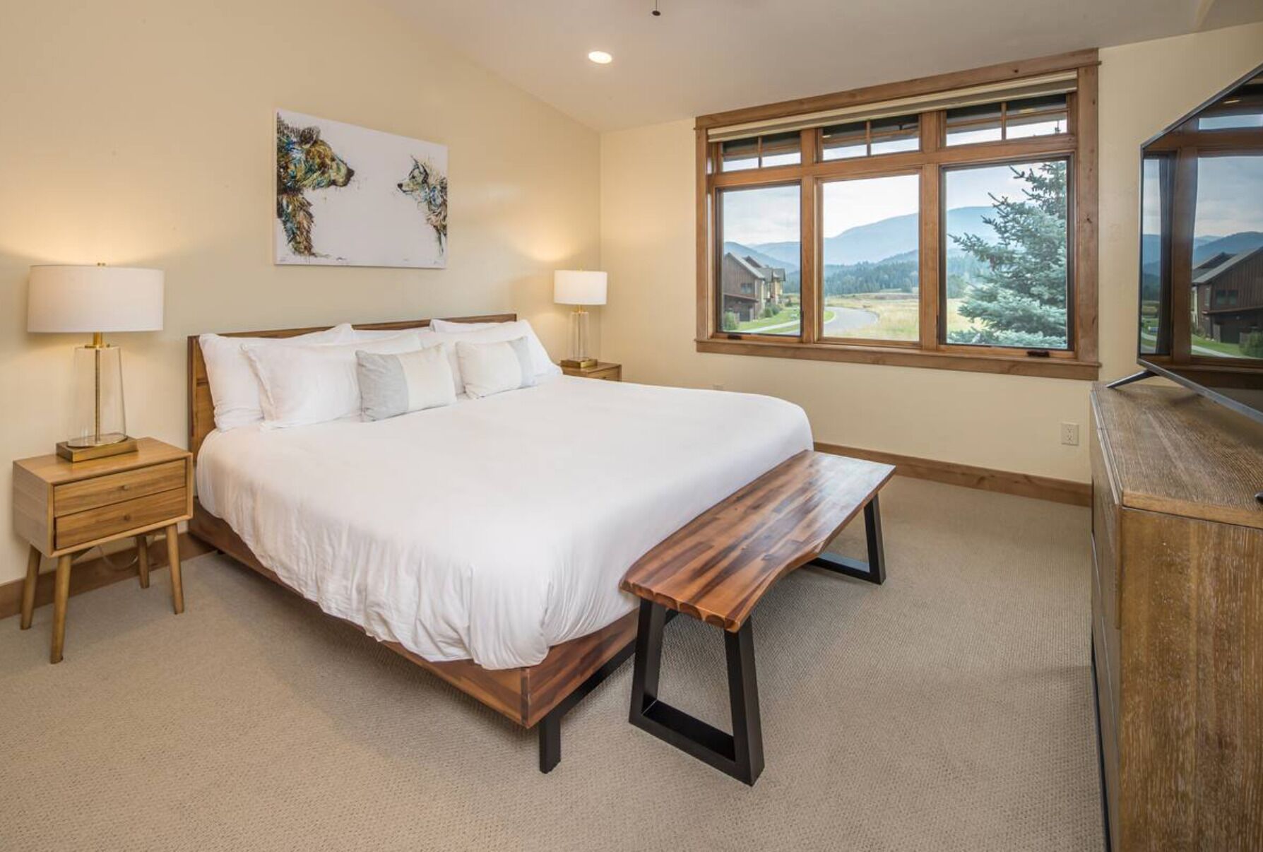 A bedroom with a large bed, bedside lamps, a bench, a painting, and a view of the outdoors.