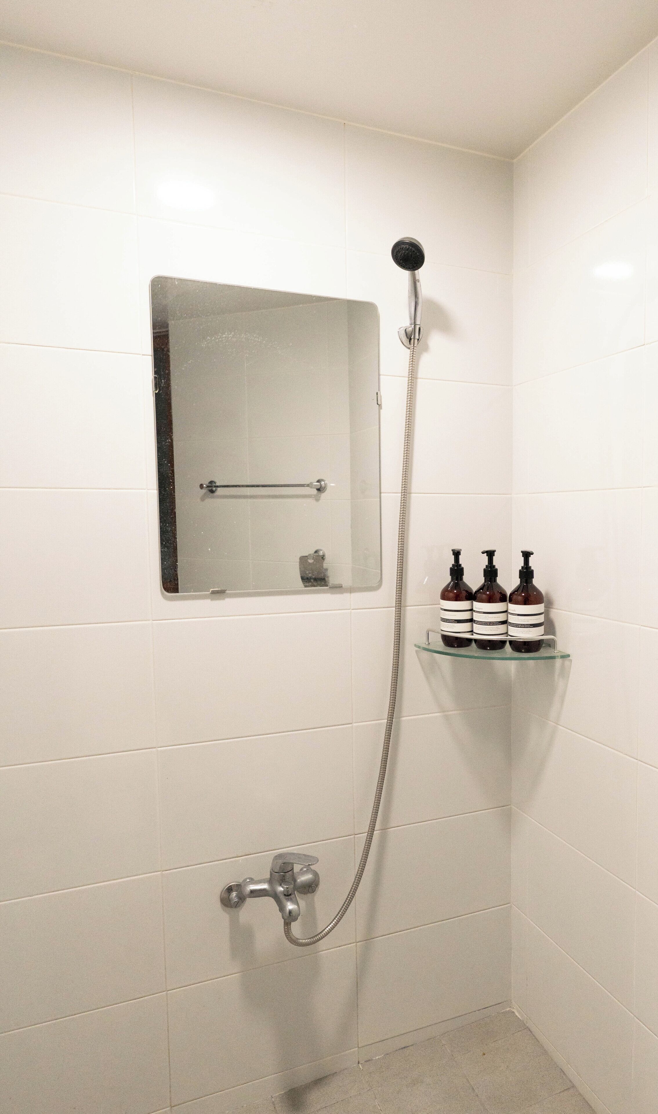 A shower with a handheld showerhead, a glass shelf with toiletries, and a small mirror on the wall.