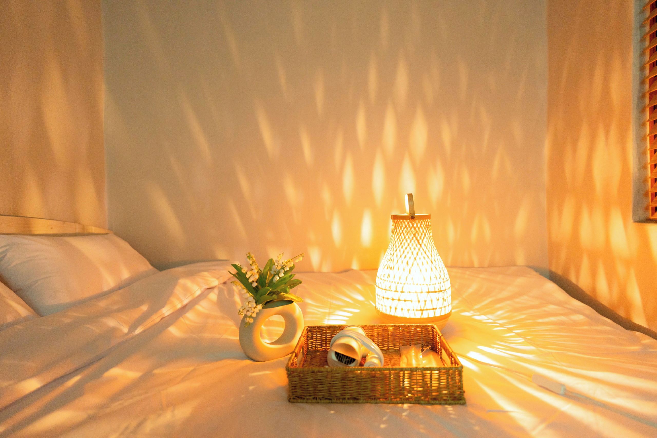 A bed with a decorative lamp, a small vase with flowers, and a wicker basket with items on a white bedspread.