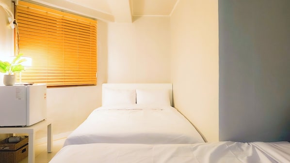 Quarto de hotel pequeno e bem organizado, com uma cama de solteiro, mesa de cabeceira com abajur e janela com persianas.