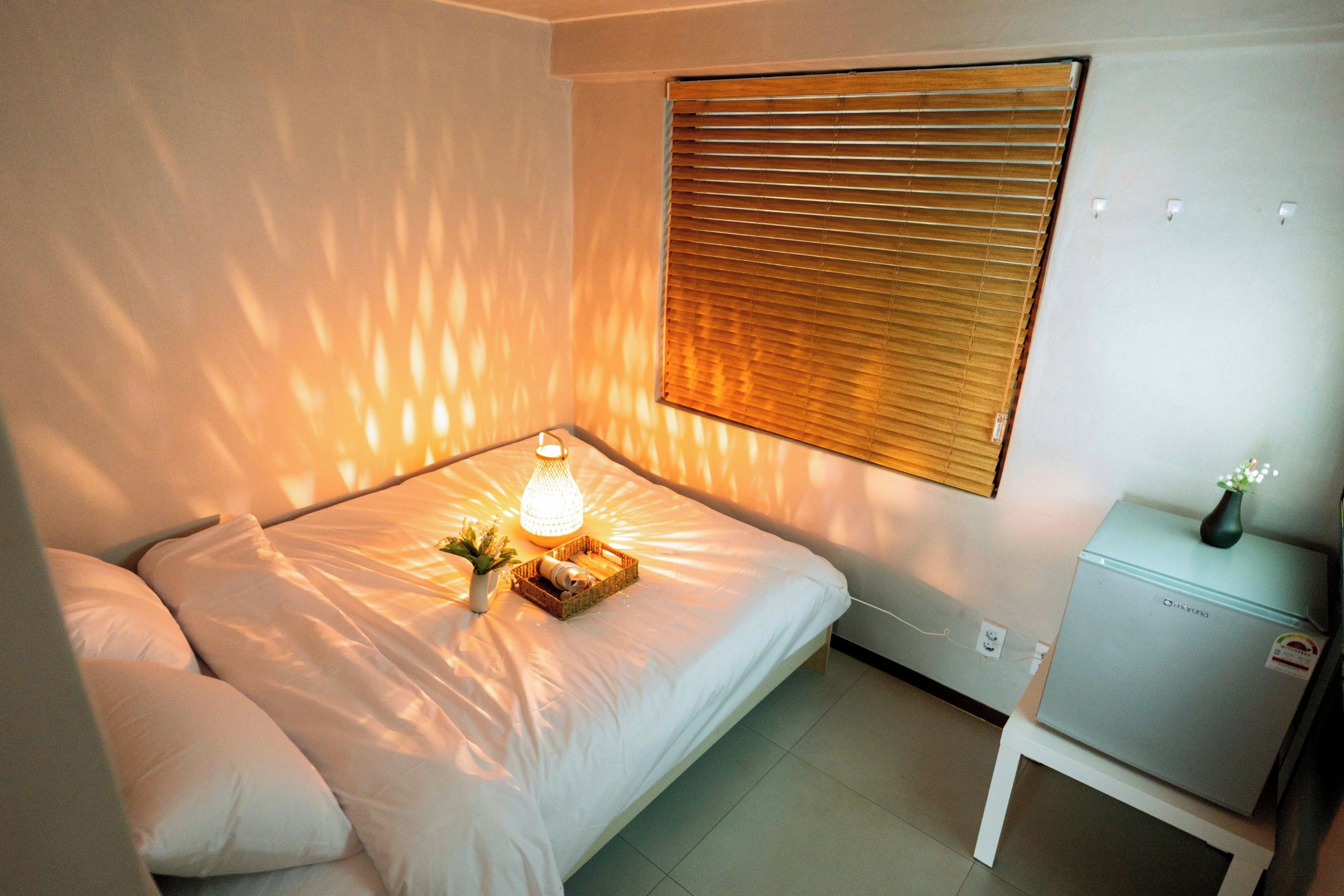 A bedroom with a bed, a bedside table, a lamp, and a window with blinds.