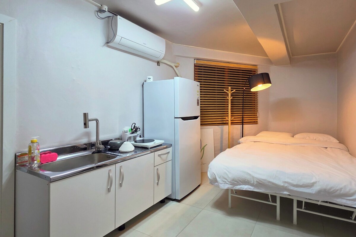 Quarto compacto com cozinha americana, uma cama, abajur e janela com persianas.