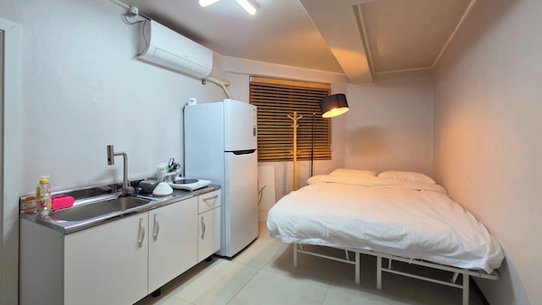 Una habitación compacta con kitchenette, cama, lámpara de noche y ventana con persianas.