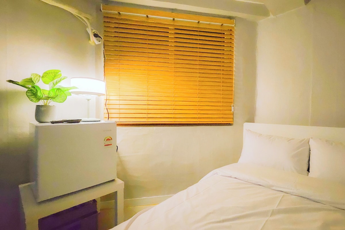 Quarto pequeno e compacto com cama, mesa de cabeceira, abajur e janela com persianas.