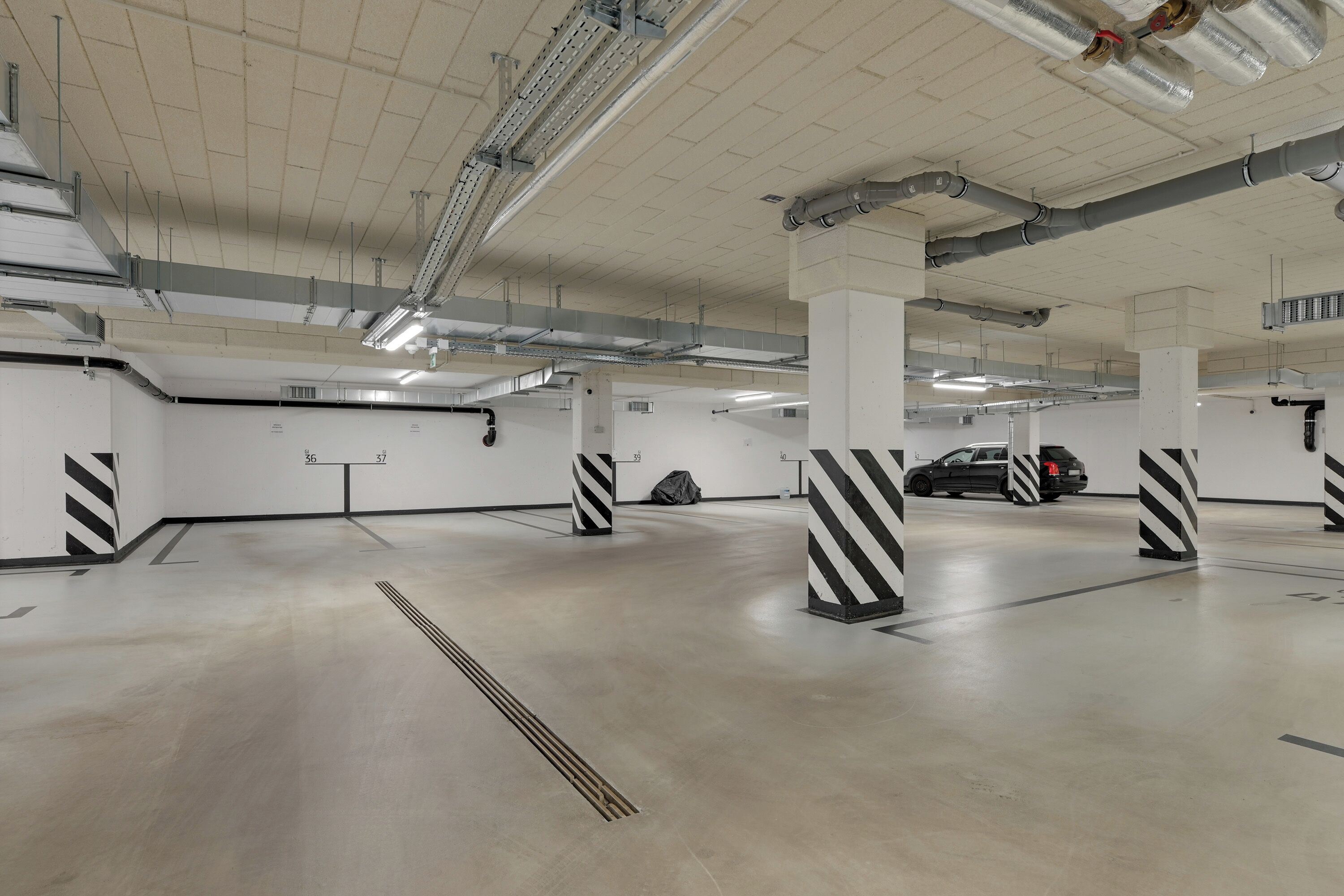 Un garaje para estacionar con pilares blancos y bases a rayas negras y blancas, pisos de concreto e iluminación colgante.