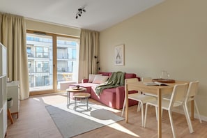Un soggiorno moderno con un divano, un tavolo da pranzo e sedie. C'è un'ampia finestra con tende, un quadro appeso al muro e vista sugli edifici esterni.