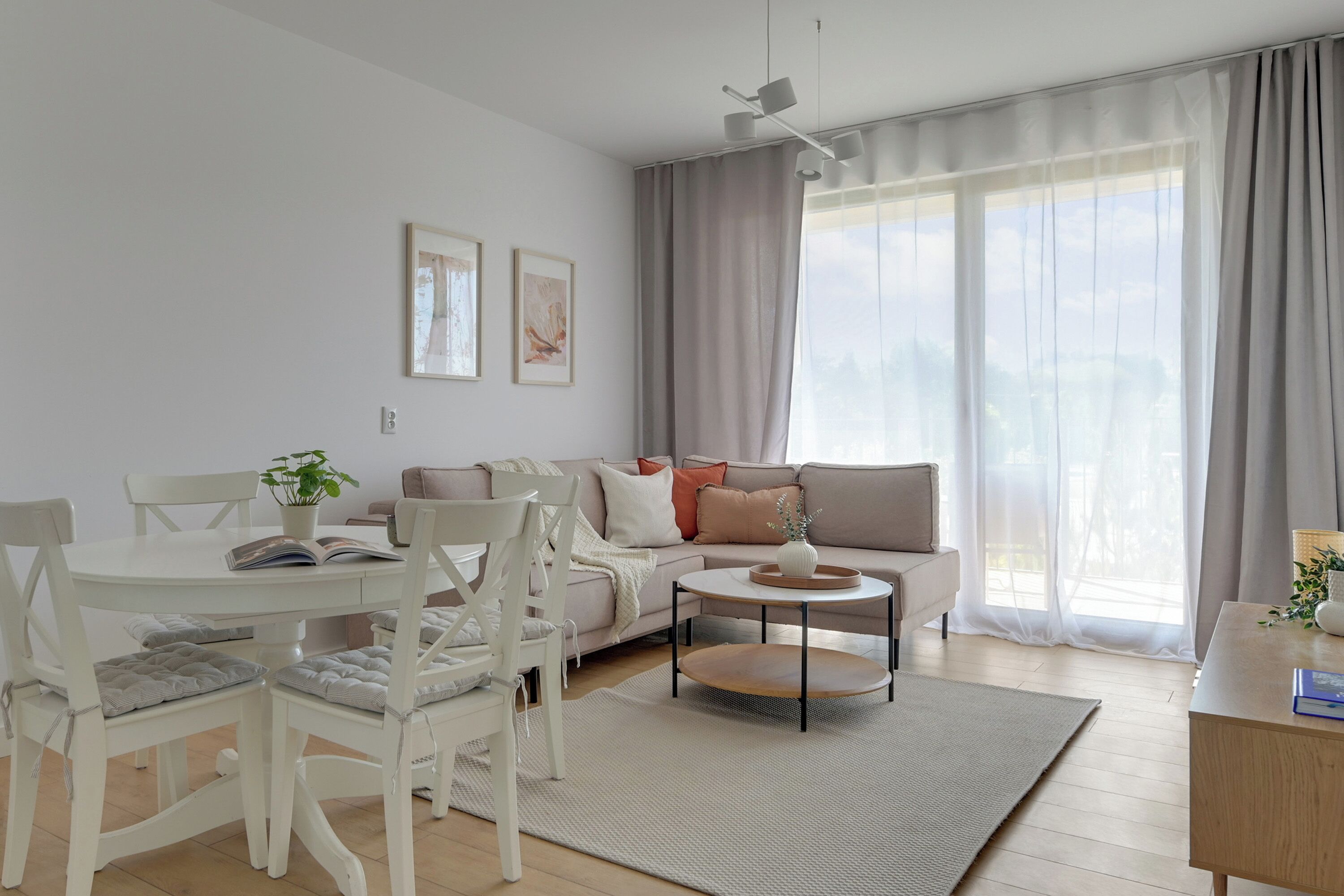 Ein modernes Wohnzimmer mit einer Couch, einem Esstisch mit Stühlen und einem Couchtisch. Es gibt ein Fenster mit Vorhängen, einen Deckenventilator und zwei gerahmte Bilder an der Wand.