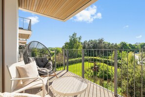 A balcony with a wicker chair and a table, overlooking a well-maintained garden and a distant building. - Slow Seaside Brzeźno by Lion Apartments (Gdańsk)