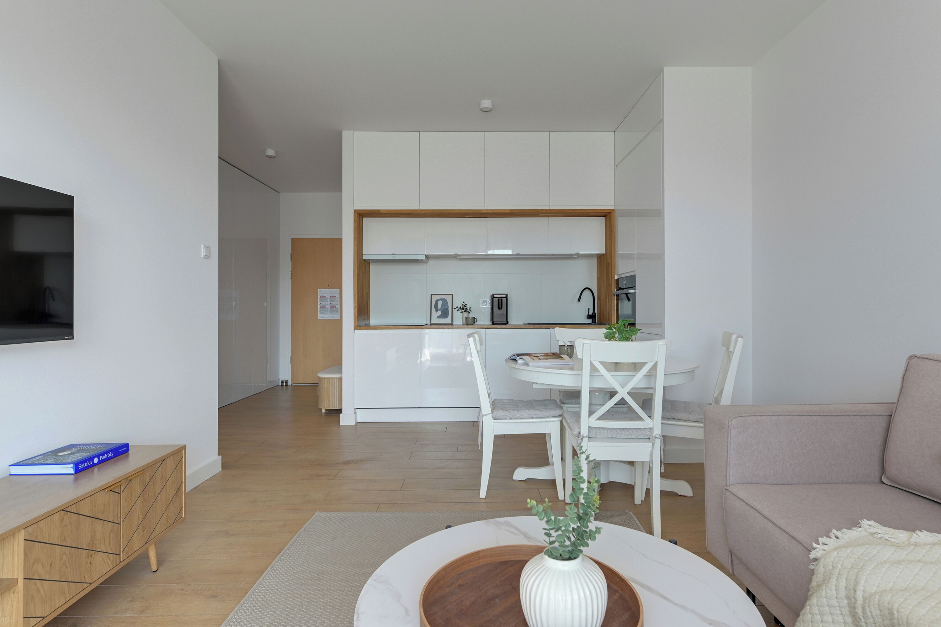 Uma sala de estar moderna com uma TV de tela plana, uma mesinha de madeira, um sofá bege e um espaço de jantar com mesa e cadeiras brancas.