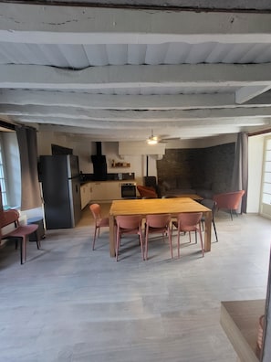 A spacious dining area with a wooden table and chairs, a kitchen in the background, and a ceiling with exposed beams.