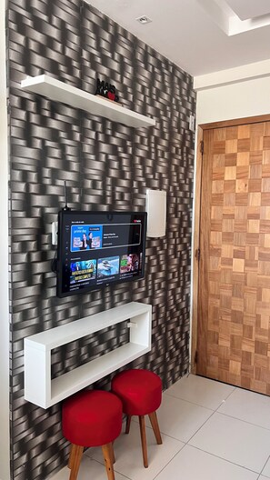 A modern interior with a flat-screen TV mounted on a textured wall, white shelving units, and a wooden door. - Apto 303 2 Qtos 15 min do Centro (Belém)