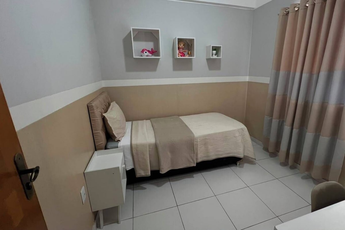 A small, single-bed room with a bedside table, a shelf with decorative items, and a window with curtains.
