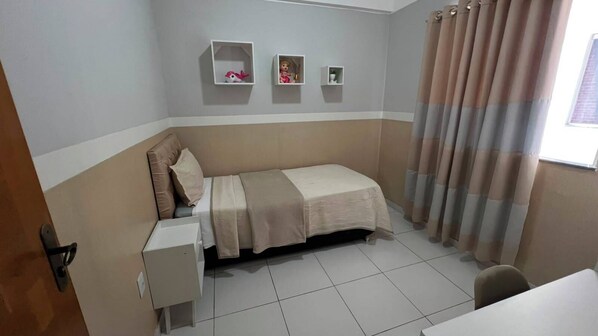 A small, single-bed room with a bedside table, a shelf with decorative items, and a window with curtains. - Apto 303 2 Qtos 15 min do Centro (Belém)
