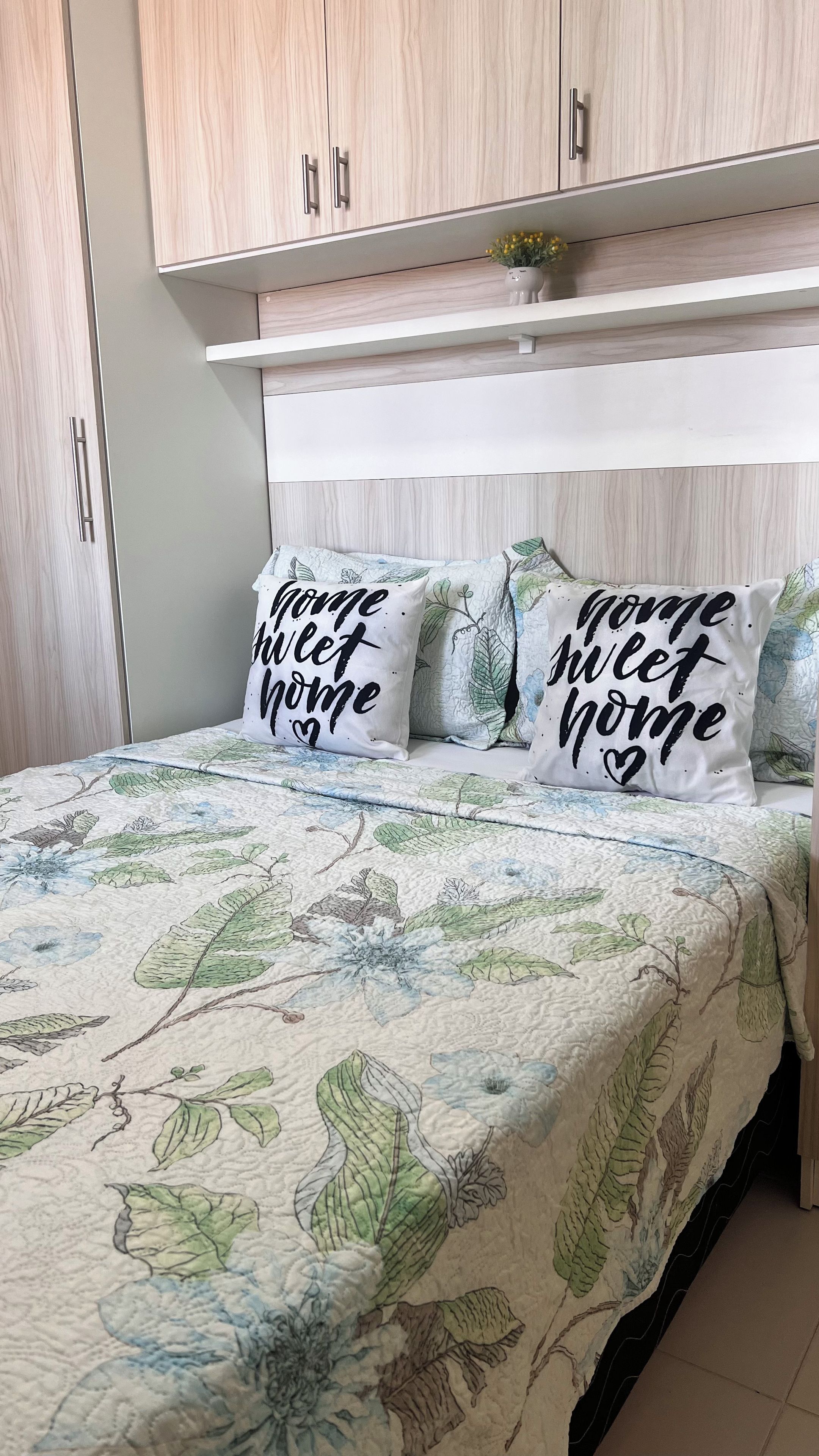 A bedroom with a bed, two pillows with 'Home sweet home' text, and a floral-patterned bedspread.