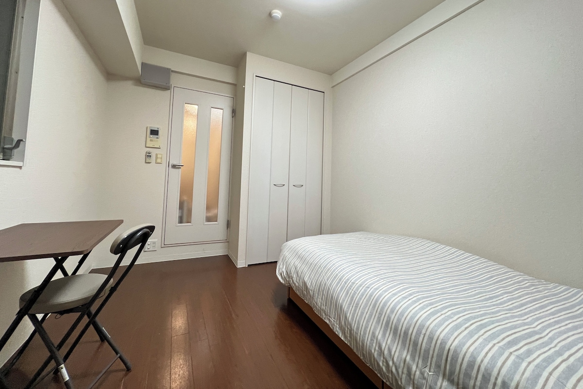 Ein kleiner, kompakter Raum mit einem Einzelbett, einem Schreibtisch und einem Stuhl. Eine Tür mit Milchglasfenster und ein kleines Fenster sind vorhanden.