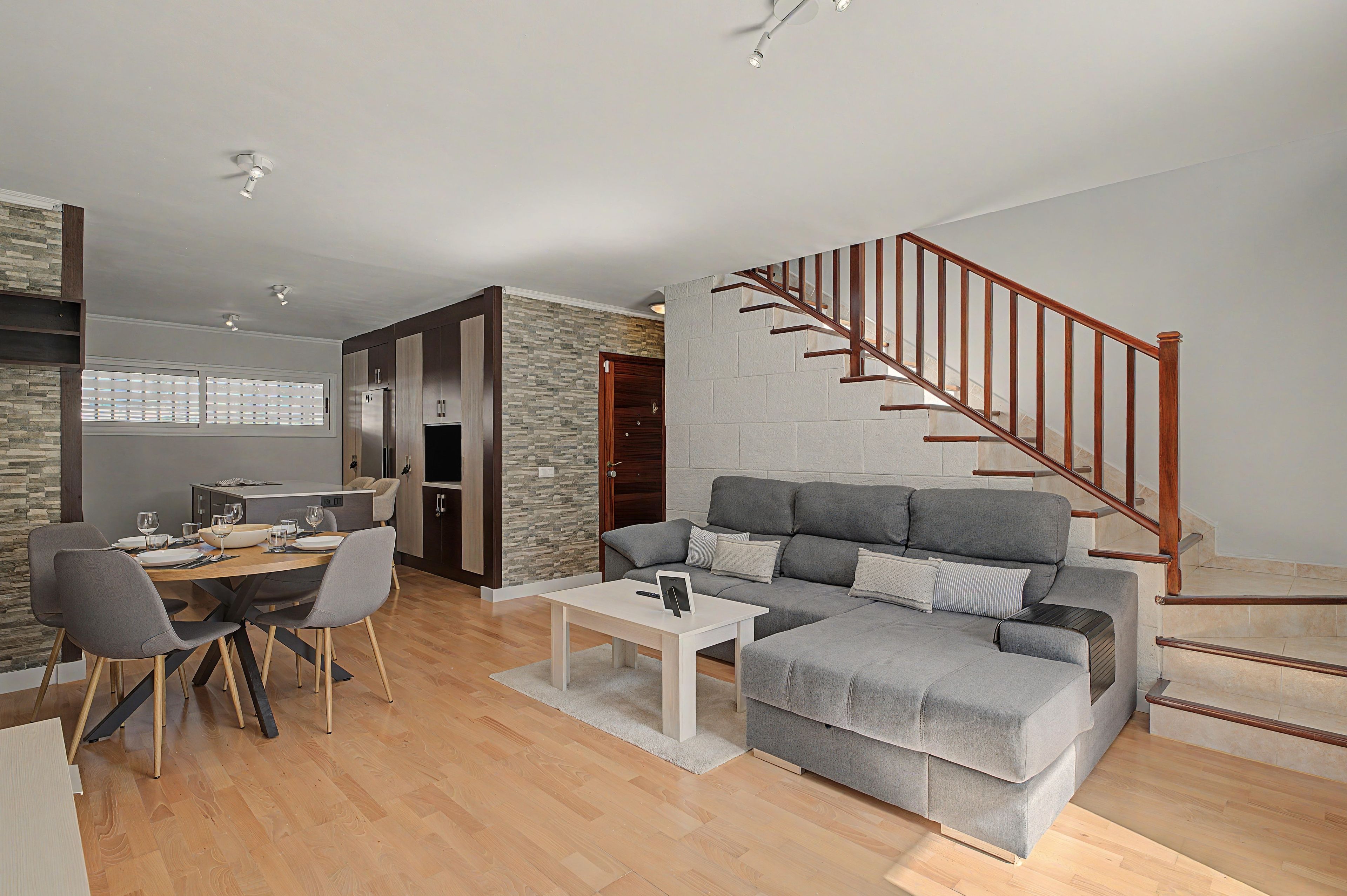 Un salon moderne avec un canapé gris, une table à manger et des chaises. Un escalier avec des rampes en bois mène à un niveau supérieur.