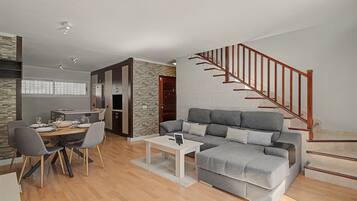 Un salon moderne avec un canapé gris, une table à manger et des chaises. Un escalier avec des rampes en bois mène à un niveau supérieur.