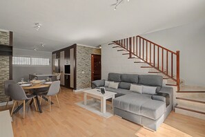 En moderne stue med en grå sofa, spisebord og stole. En trappe med trægelændere fører til en etage ovenover.