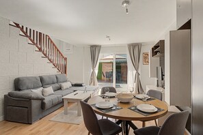 Una sala moderna con un sofá gris, una escalera de madera, un área de comedor con una mesa redonda y vistas al exterior.