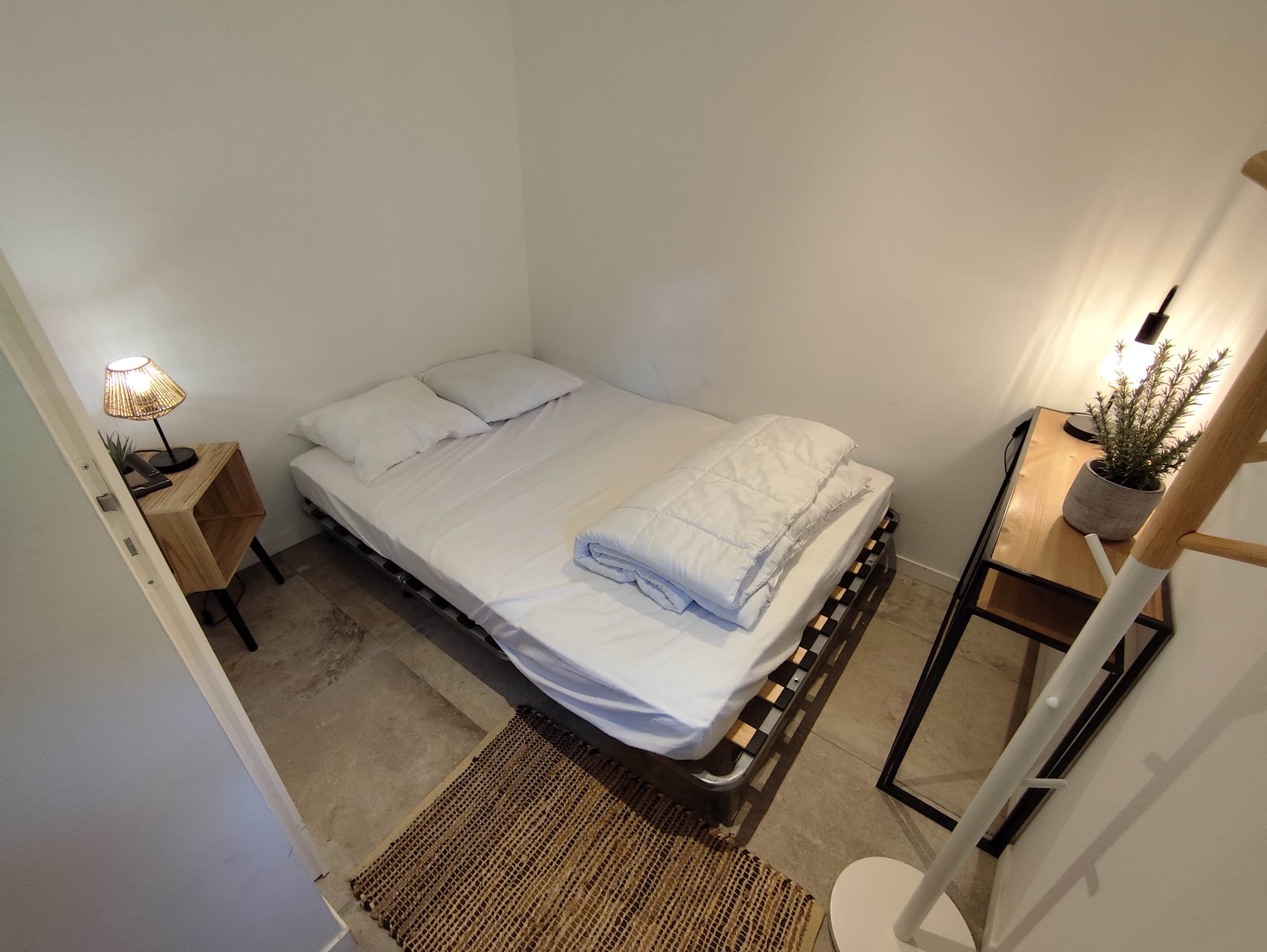 Une petite chambre avec un lit simple, une tête de lit en bois, une table de chevet avec une lampe et une plante en pot posée sur une étagère en bois.