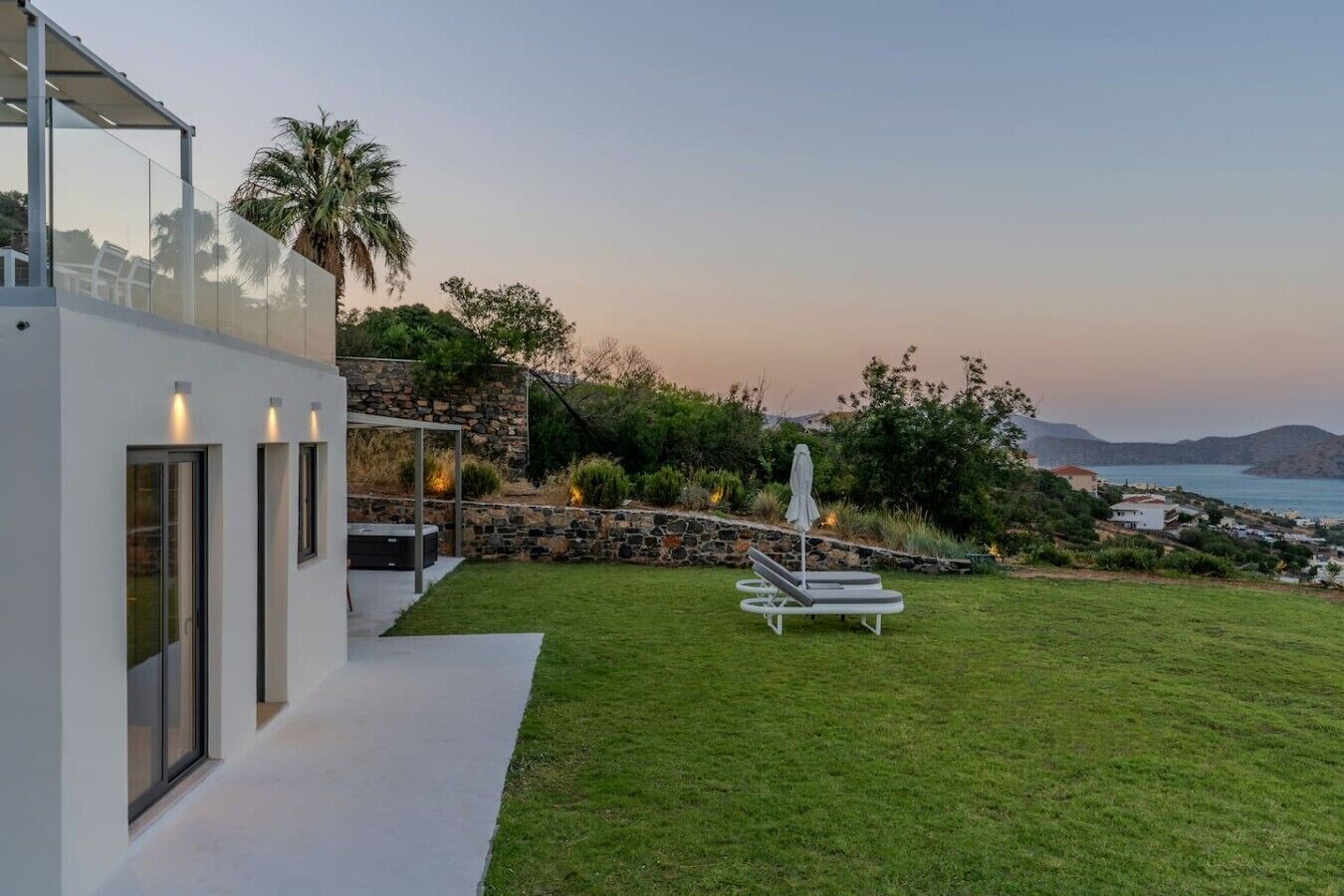 Una casa moderna con fachada de vidrio, un muro de contención de piedra, un césped y vista al mar.