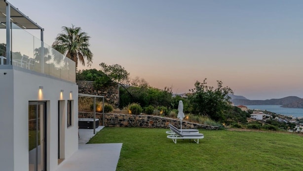 Una casa moderna con facciata in vetro, un muro di sostegno in pietra, un prato e vista sul mare.