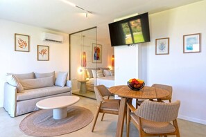 Ein modernes Wohnzimmer mit einer Couch, einem runden Esstisch mit Stühlen, einem flachbildfernseher an der Wand und einem kleinen Beistelltisch mit Lampe.
