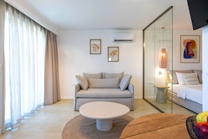 Ein modernes Hotelzimmer mit einer Couch, einem runden Couchtisch, einem Bett mit Nachtschränkchen und einem großen Spiegel.