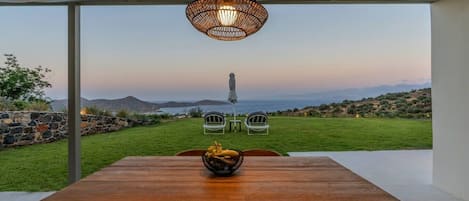 A wooden table with two chairs, a wicker pendant light, and a view of the sea and mountains.