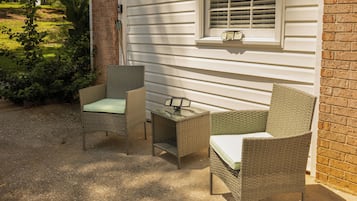 An outdoor seating area with wicker furniture and a small table, adjacent to a house with a window and a brick wall.