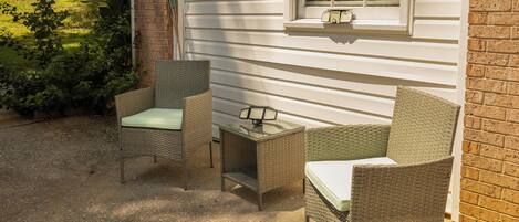 An outdoor seating area with wicker furniture and a small table, adjacent to a house with a window and a brick wall.