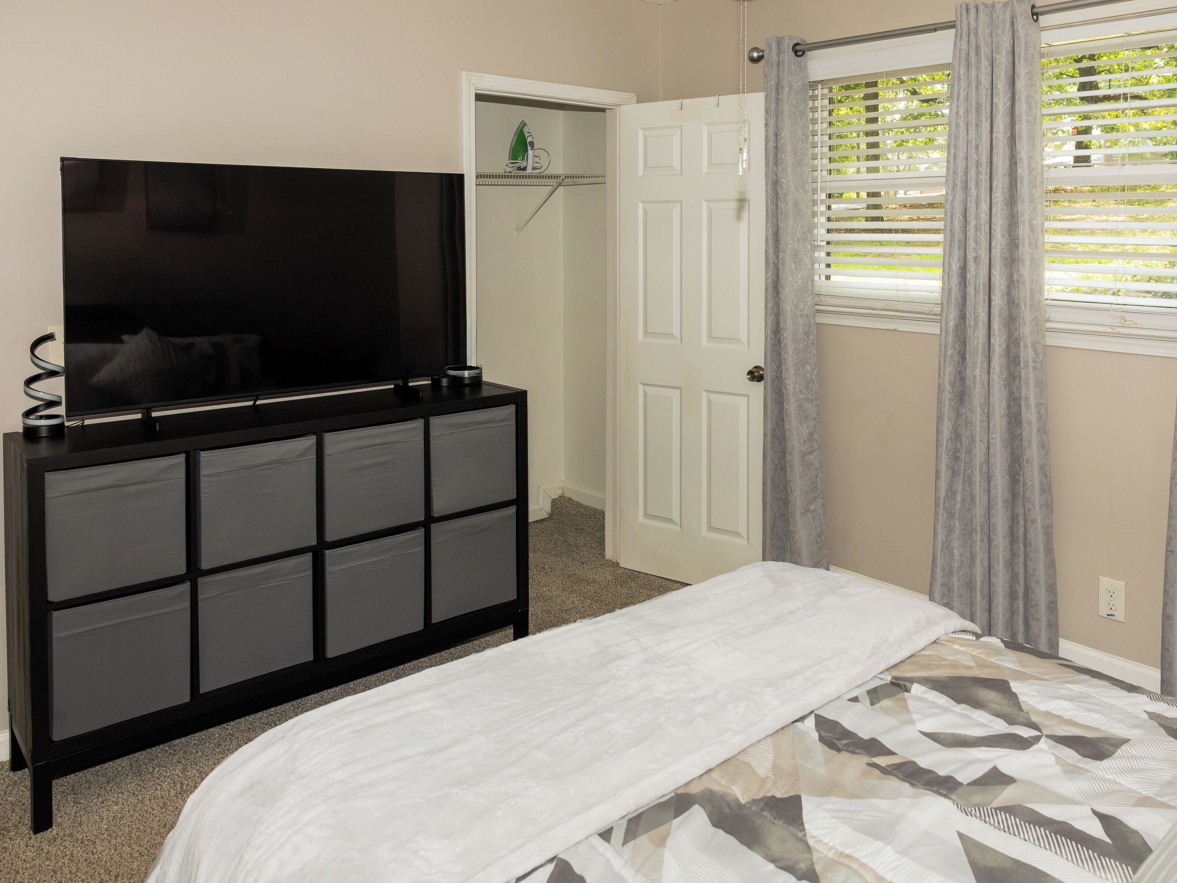 Un dormitorio con cama, televisor sobre una cómoda, puerta y ventana con cortinas.