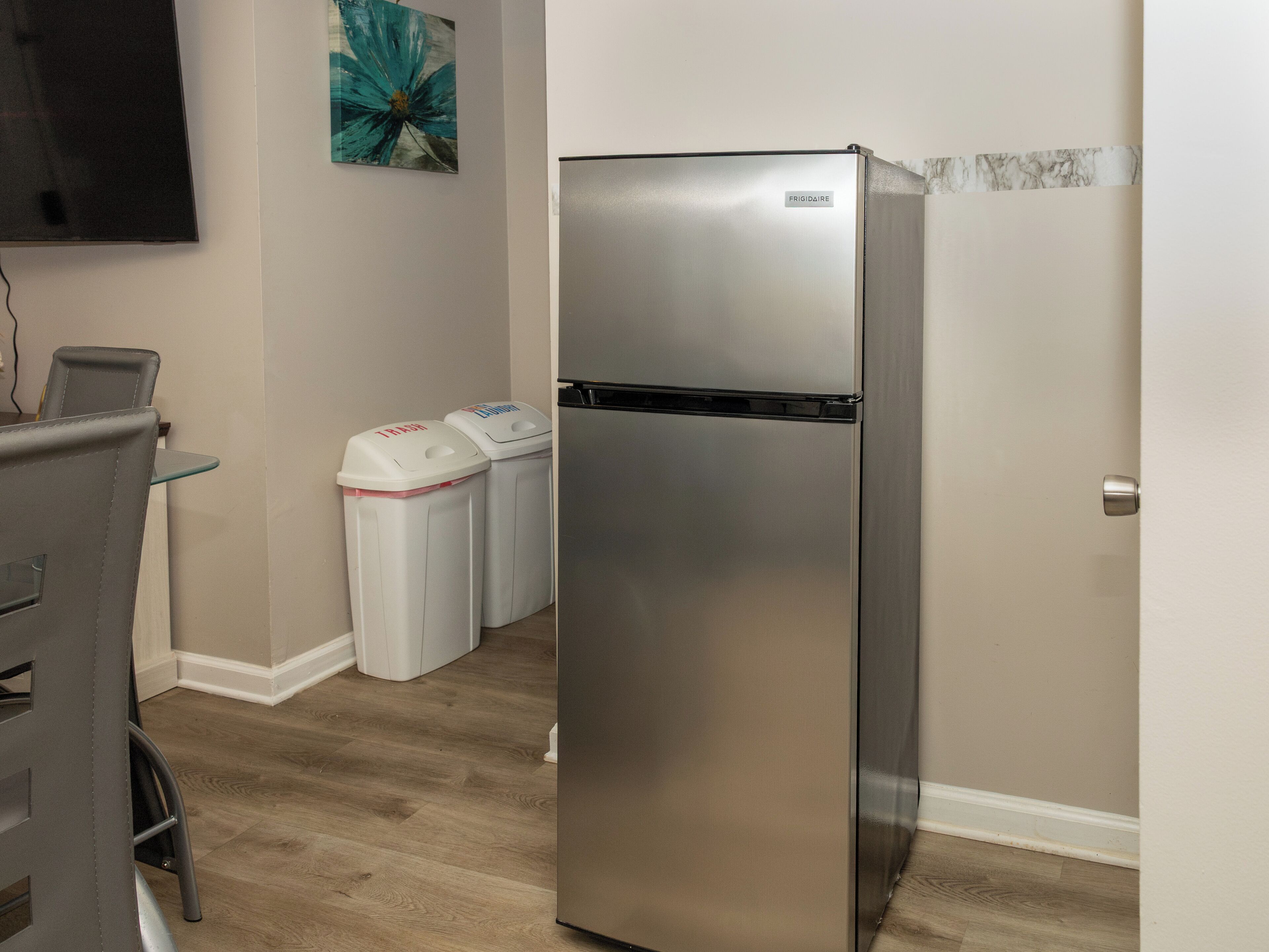 Une cuisine équipée d’un réfrigérateur en acier inoxydable, de deux poubelles, d’une table à manger avec des chaises et d’un téléviseur à écran plat fixé au mur.