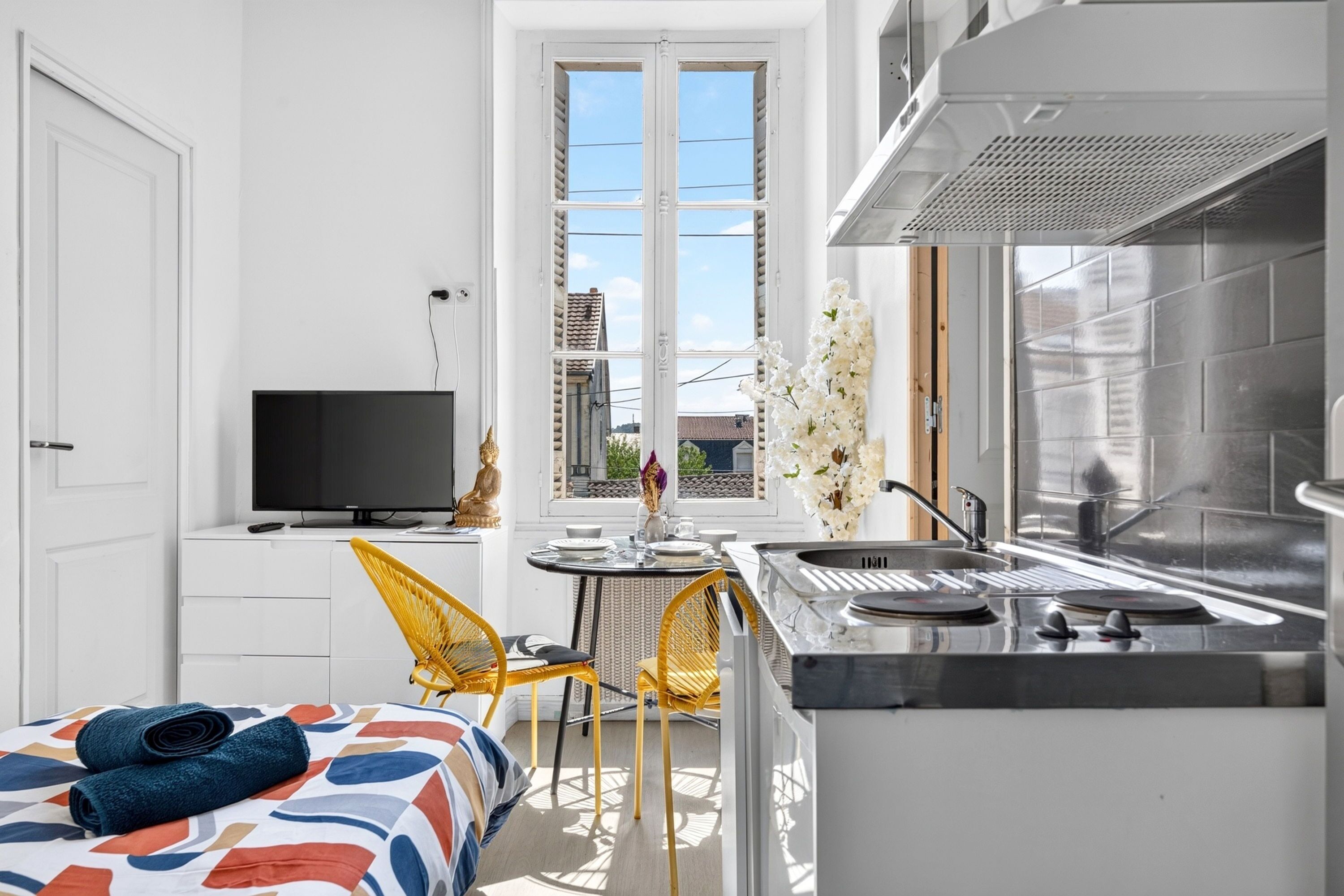 Ett kompakt hotellrum med en säng, en TV, en matplats med stolar, ett kök med diskho och ett stort fönster.