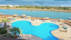 Una piscina con bordo curvo, circondata da lettini e ombrelloni, con vista su uno specchio d'acqua e edifici in lontananza.