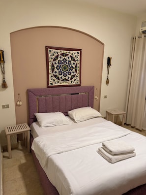 Una habitación de hotel con cama, cabecera, una obra de arte en la pared, dos mesitas de noche y una ventana con cortinas.