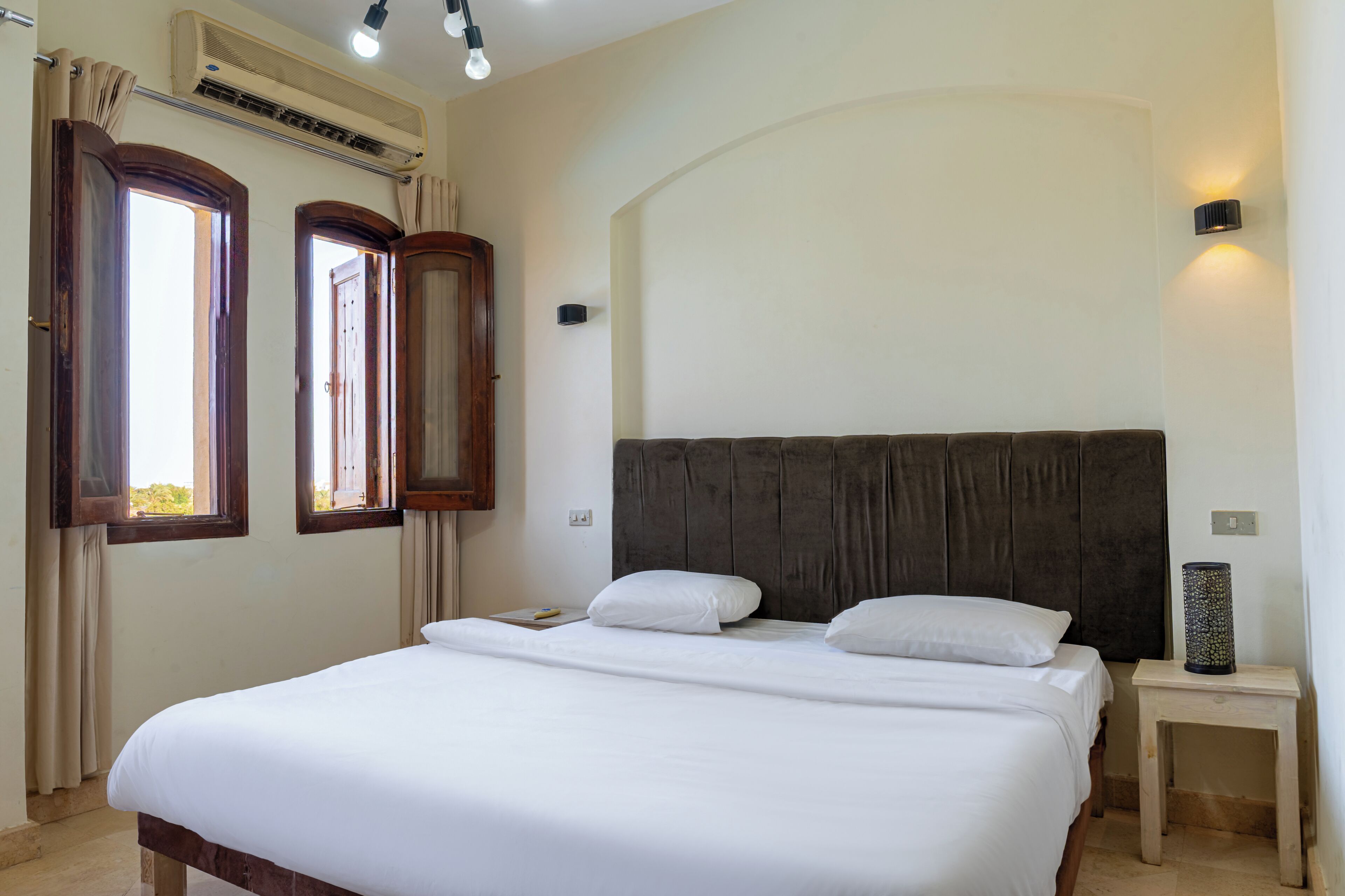A bedroom with a large bed, two windows, an air conditioning unit, a bedside lamp, and a nightstand.