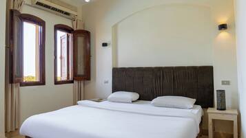 A bedroom with a large bed, two windows, an air conditioning unit, a bedside lamp, and a nightstand.
