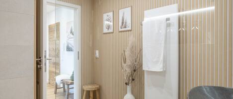 Un baño moderno con pared de paneles de madera, una toalla blanca colgada en un perchero, un jarrón grande con plantas secas y un taburete pequeño.
