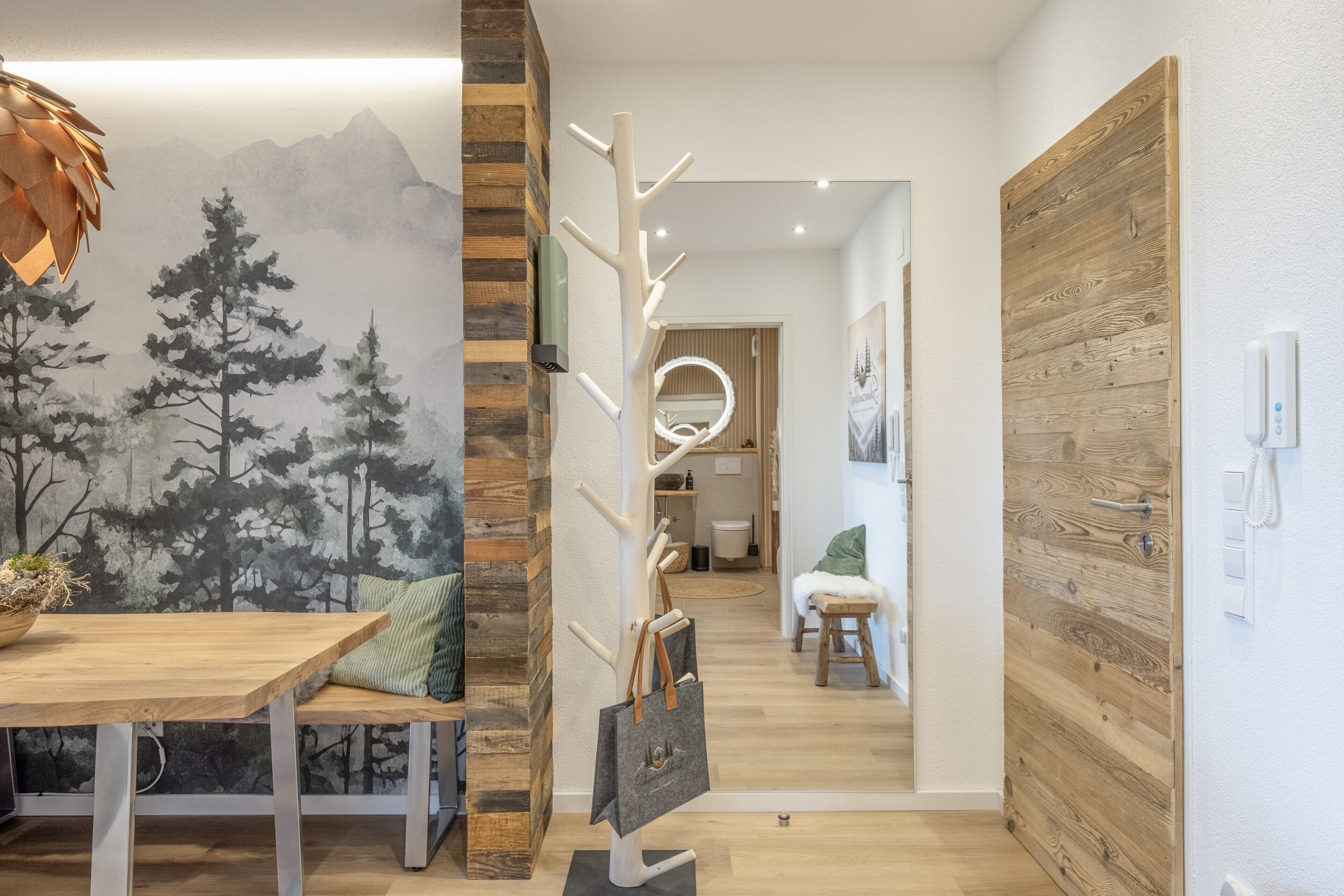 Un interno moderno con un tavolo da pranzo in legno, un appendiabiti e una parete con un murale raffigurante montagne e alberi.