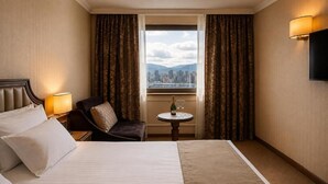 Habitación de hotel con una cama, una silla, una mesita con una botella y un vaso, y una ventana con cortinas.