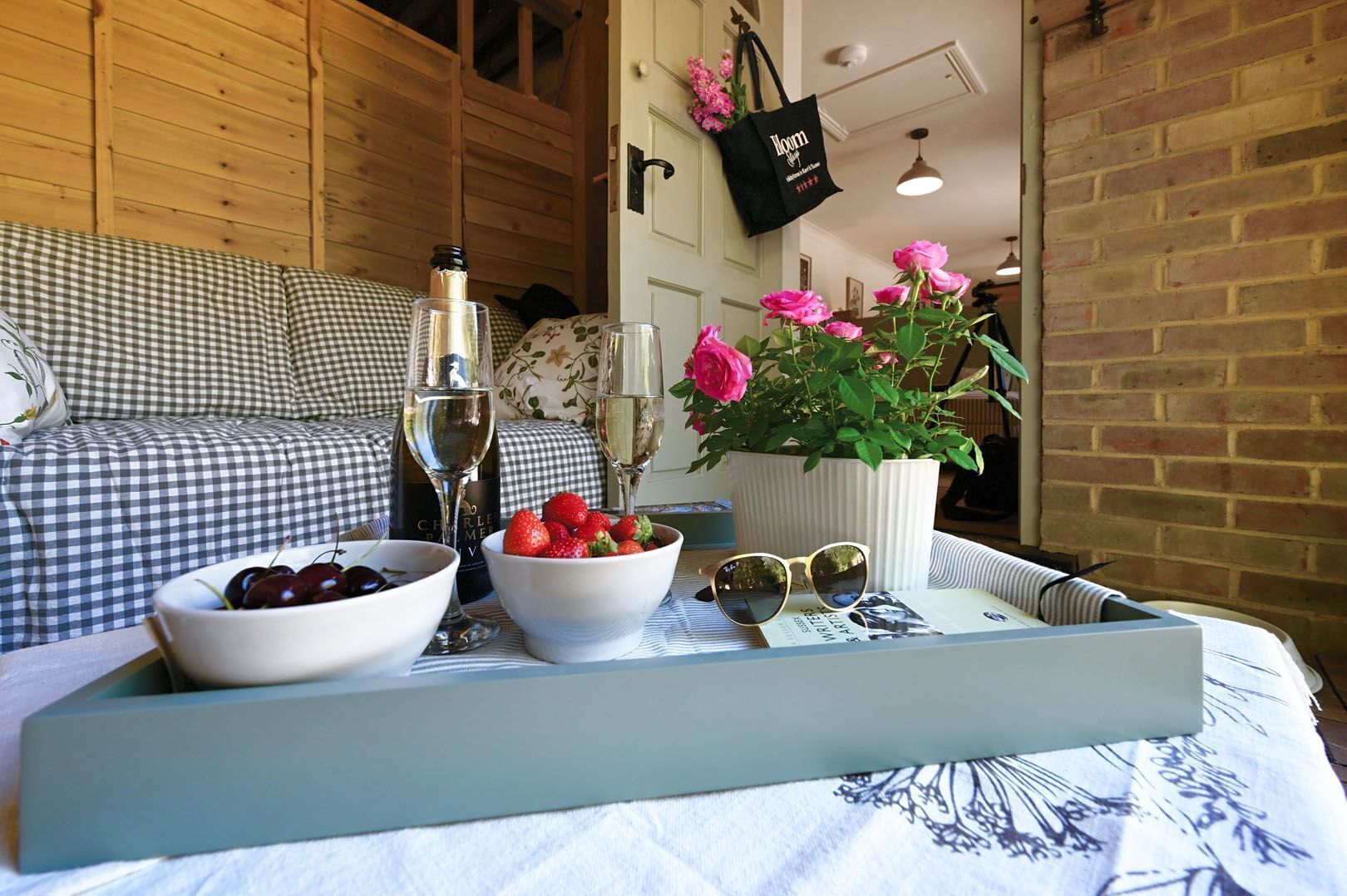 A cozy room with a checkered sofa, a tray with fruit, wine glasses, and a vase with flowers.