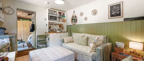 Uma sala de estar acolhedora, com um sofá, um banquinho e uma mesinha com abajur. Há uma área de cozinha com micro-ondas e pia.
