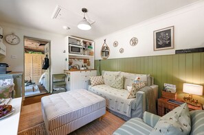 A cozy living room with a sofa, ottoman, and a small table with a lamp. There is a kitchen area with a microwave and a sink.